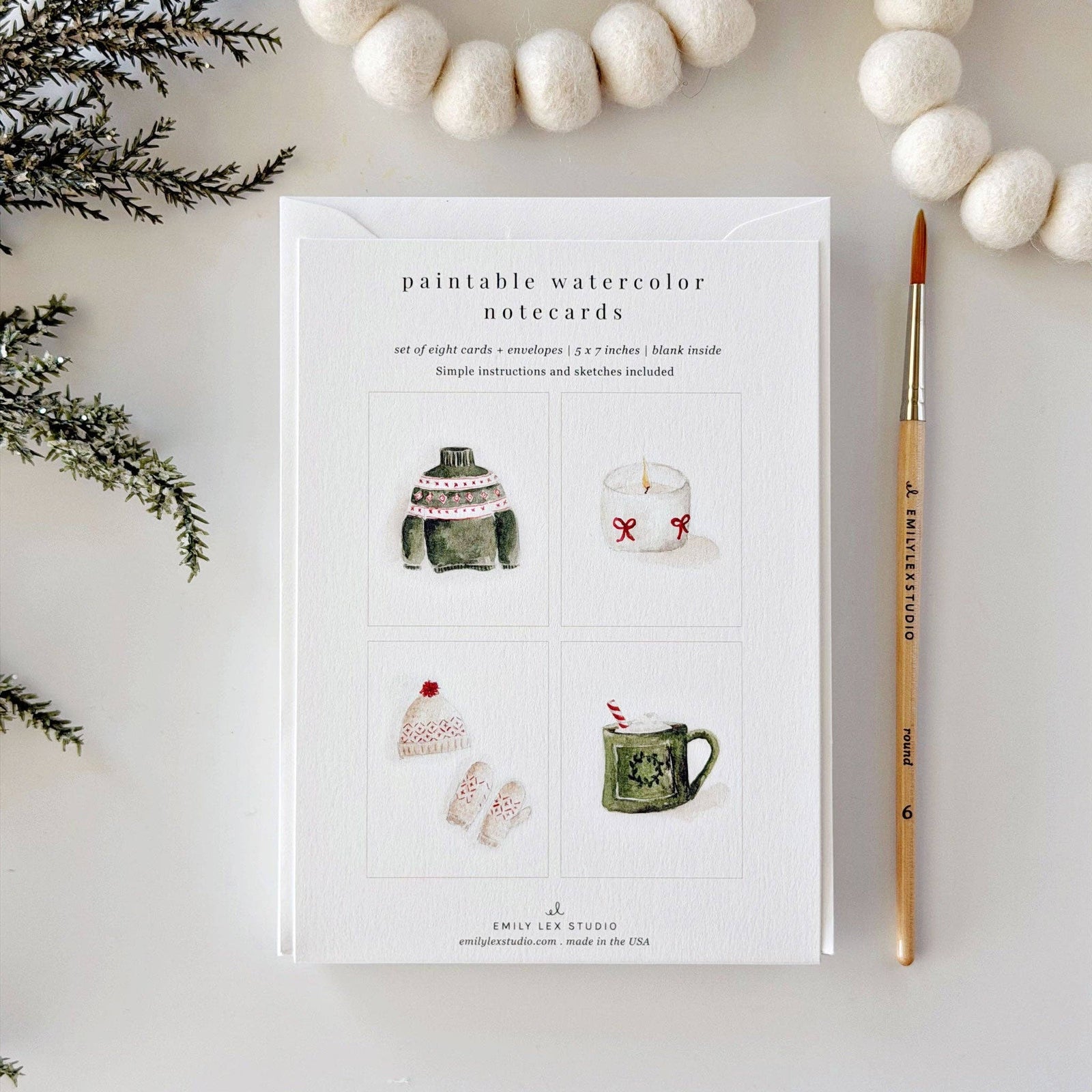 A flat-lay of a notepad card titled 'paintable watercolor notecards' showing four small watercolor illustrations, styled with white felt balls, pine branches, and a paintbrush on a light background.