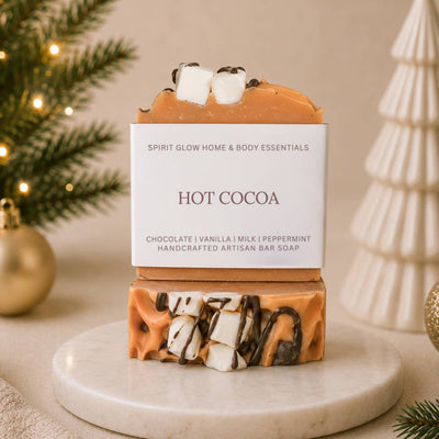 A handcrafted artisan bar soap labeled 'Hot Cocoa' styled on a marble coaster with festive holiday decorations in the background.