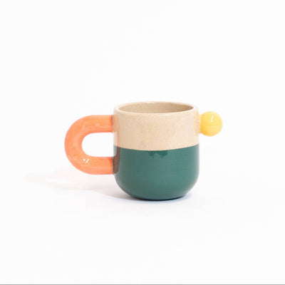 A small ceramic mug with a split design, featuring a dark teal lower half, a beige upper half, a curved orange handle, and a small yellow accent on the side, set against a plain white background.