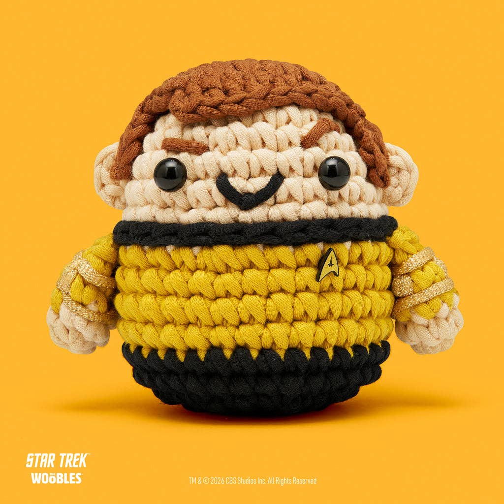 A crocheted plush toy resembling a Star Trek character, with cream-colored yarn for skin and hair, brown yarn hair, a yellow and black uniform, and a small gold delta insignia on the chest, set against a solid orange background. Star Trek branding is visible in the bottom left corner.
