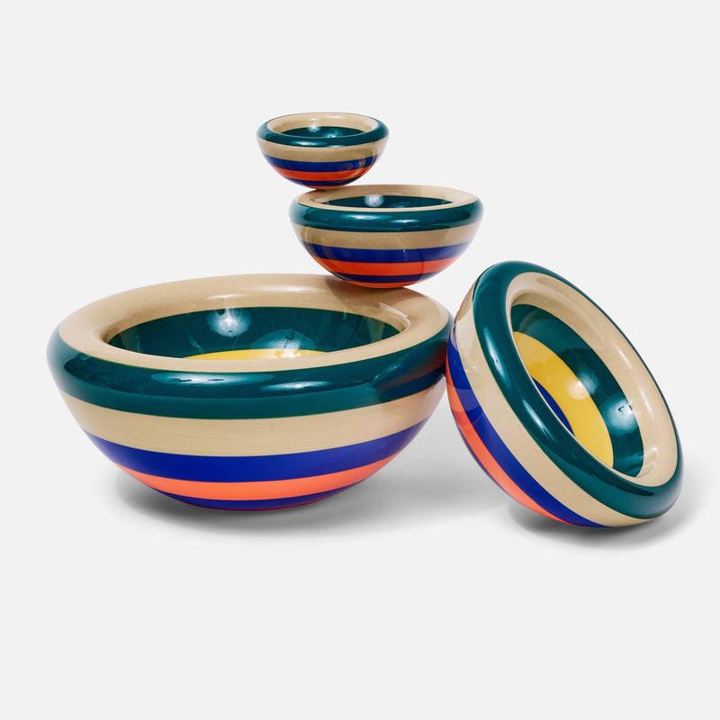 A person's hands holding a round, shallow dish with concentric target-style stripes in dark teal, cream, yellow, and blue against a plain white background.