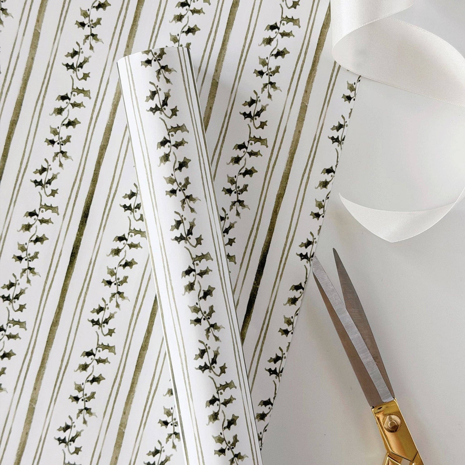 A close-up of patterned wrapping paper with vertical olive green stripes and ivy vines on a white background, with a pair of gold-handled scissors and a rolled sheet of white paper on a light surface.