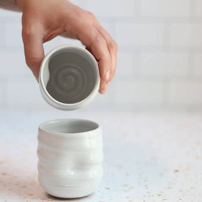 Hand holding a small ceramic cup over another similar cup on a light background