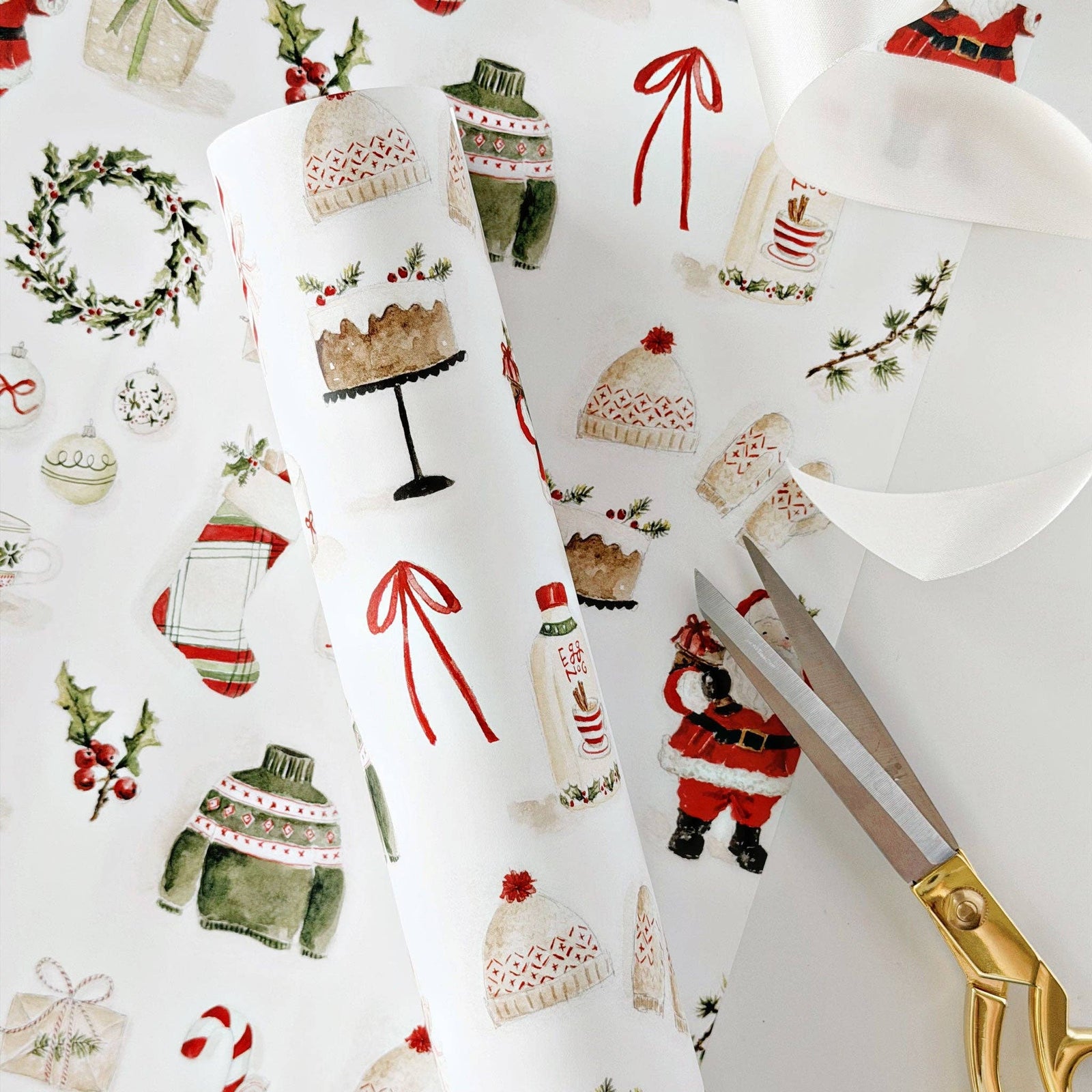 A roll of Christmas-themed wrapping paper with a festive watercolor print, shown alongside gold-handled scissors and a matching flat sheet of the paper on a white background.