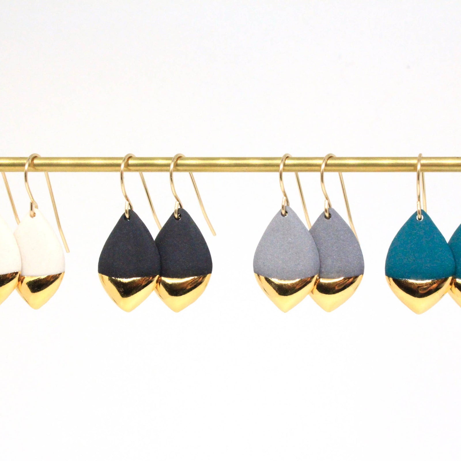 Four pairs of teardrop-shaped dangle earrings hanging from a gold rod against a white background. The earrings have a two-tone design with a matte top half and a polished metallic bottom half.