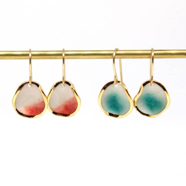 Two pairs of gold hoop earrings with irregular teardrop-shaped white enamel pendants are displayed on a gold bar. The left pair is solid white, the middle pair has a soft red gradient at the bottom, and the right pair has a teal-blue gradient at the bottom.