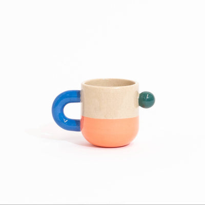 A small ceramic mug with a split design, featuring a beige upper half, a coral lower half, a blue handle, and a small green accent on the side, set against a plain white background.