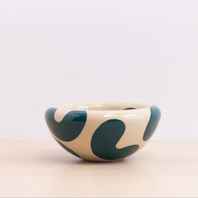 A small ceramic bowl with an organic wavy pattern in teal on a warm cream-colored glaze, set on a light wooden surface against a white background.
