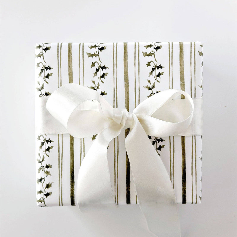 A close-up of patterned wrapping paper with vertical olive green stripes and ivy vines on a white background, with a pair of gold-handled scissors and a rolled sheet of white paper on a light surface.