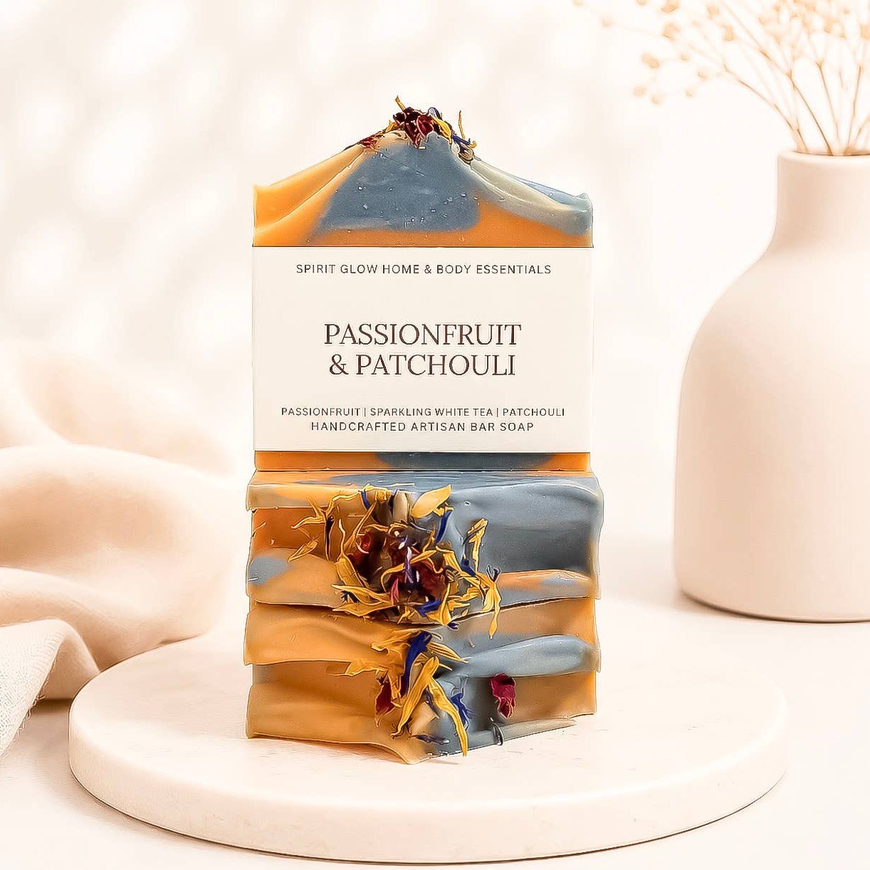 A handmade artisan bar soap labeled 'PASSIONFRUIT & PATCHOULI' by Spirit Glow Home & Body Essentials, stacked on a white marble tray with dried botanicals, set against a soft beige background with a ceramic vase.
