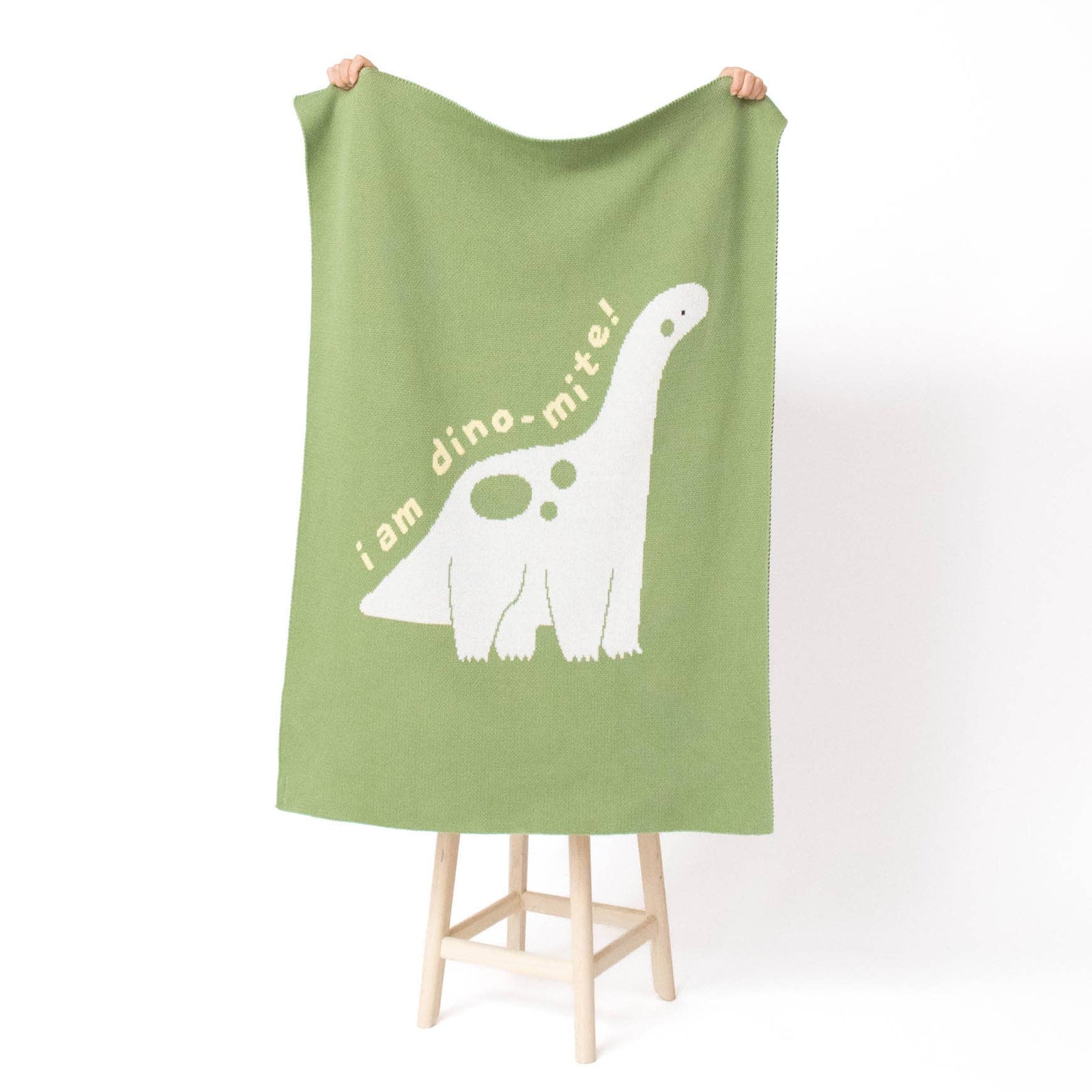 A sage green blanket with a white long-necked dinosaur and the text 'i am dino, mite!' is held up by two hands on a small wooden stool against a white background.
