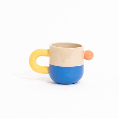 A small ceramic mug with a split design, featuring a beige top half, a blue bottom half, a yellow handle, and a small orange accent on the side, set against a plain white background.