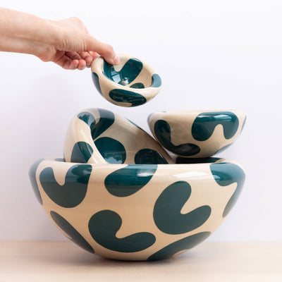 A hand holds a small matching bowl above a set of nested ceramic bowls with a cream background and dark teal abstract wavy spots, photographed against a clean white wall.