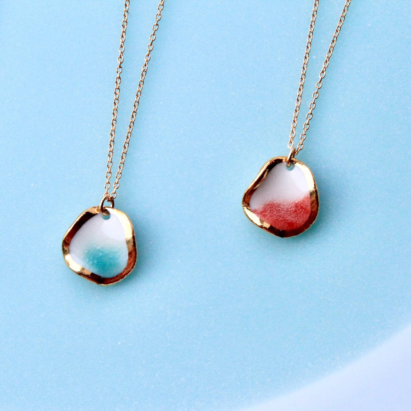 Two gold-tone chain necklaces with irregular organic-shaped pendants are arranged on a light blue background. 