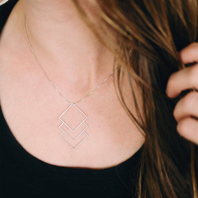 A close-up lifestyle shot of a woman wearing a delicate silver necklace with three nested, open geometric diamond shapes. The necklace is shown against her skin and a black top, with her brown hair framing the side of the image.