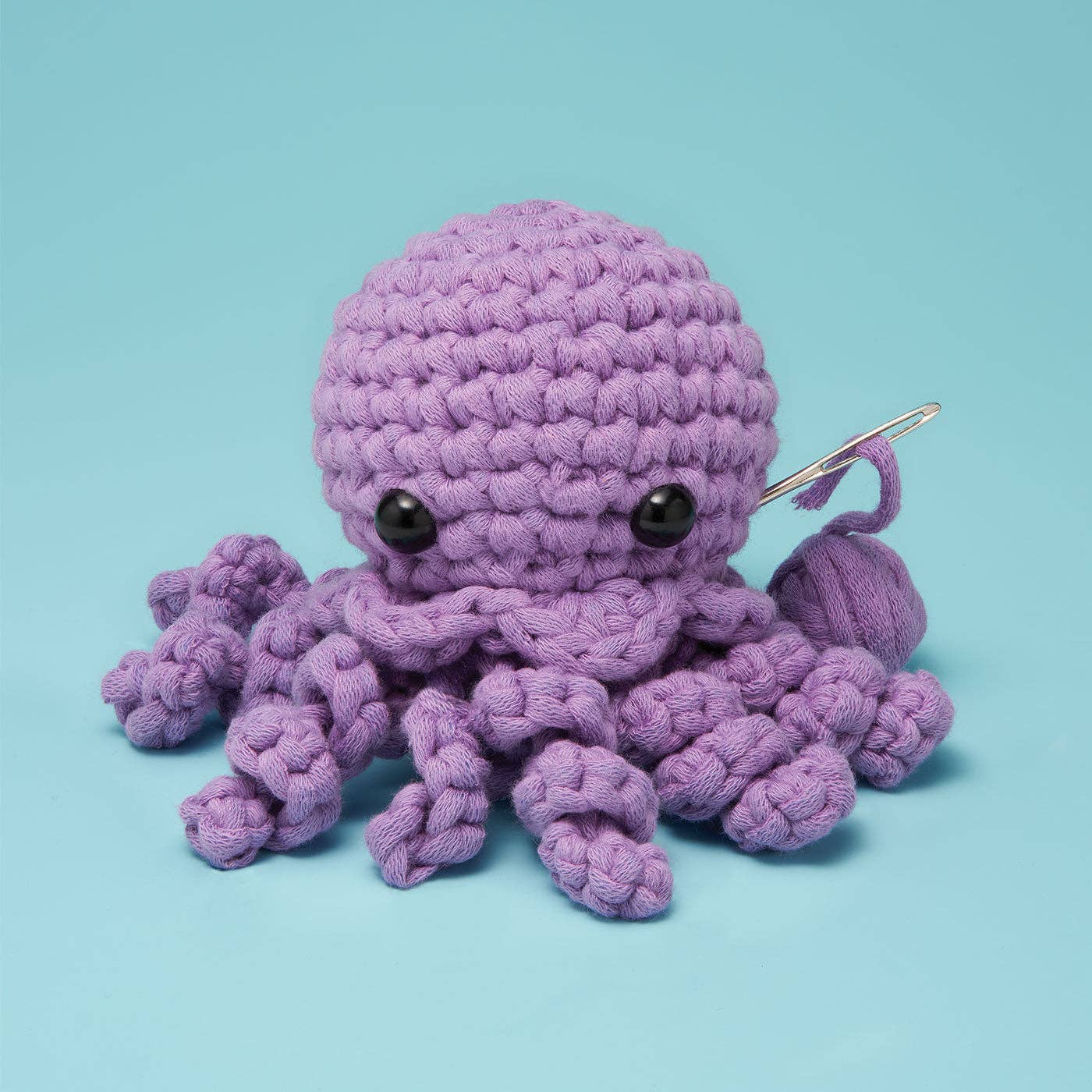 A purple crocheted octopus toy with black bead eyes and a silver crochet hook attached to its tail, set against a solid light blue background.