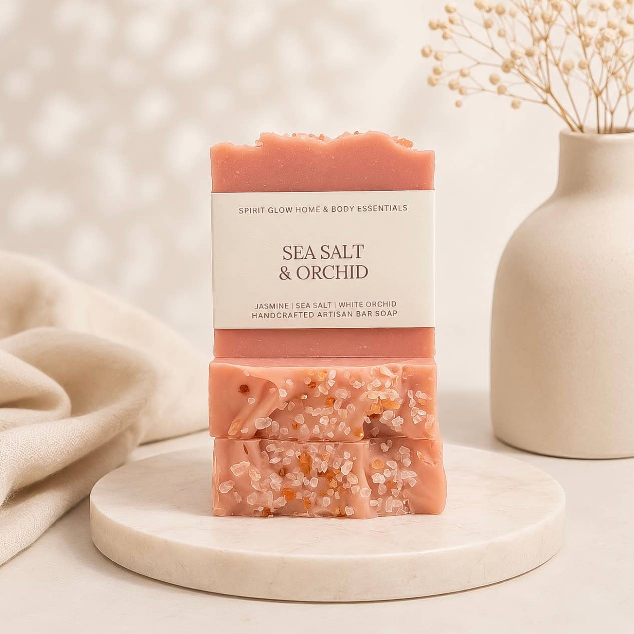 A bar of handmade body soap labeled 'SEA SALT & ORCHID' by Spirit Glow Home & Body Essentials, displayed on a round white marble tray. The soap has a dusty pink top and bottom half with a white center band, and is styled with a beige fabric and a ceramic vase in the background.