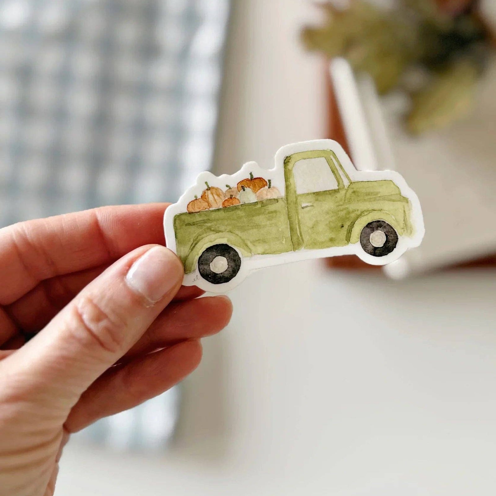 A hand holding a sticker of a vintage green truck with pumpkins on a light blurry background