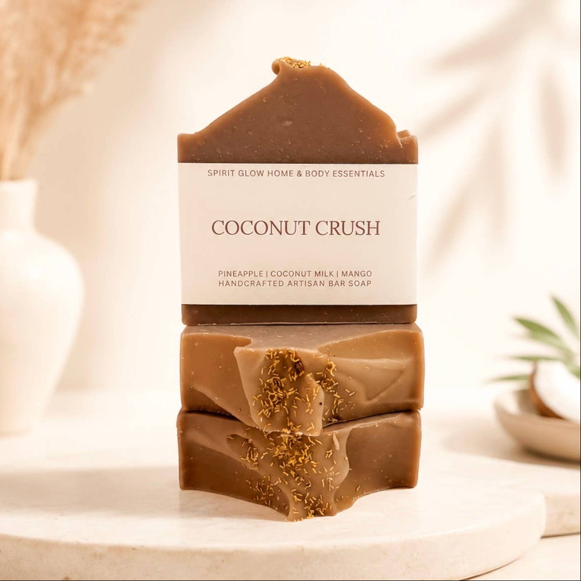 Three stacked artisan bar soaps labeled 'COCONUT CRUSH' by Spirit Glow Home & Body Essentials, displayed on a round marble tray with dried grass and a small plant in the background.