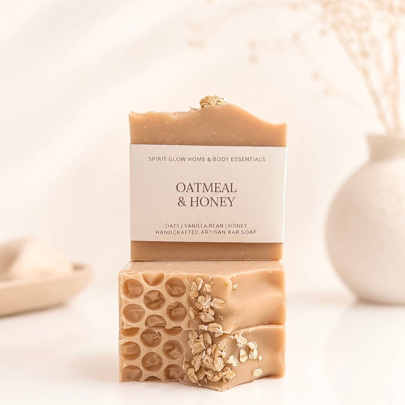 A bar of oatmeal and honey artisan soap displayed in two stacked pieces to show its honeycomb texture and oat topping, with a paper label on a warm, softly lit background.