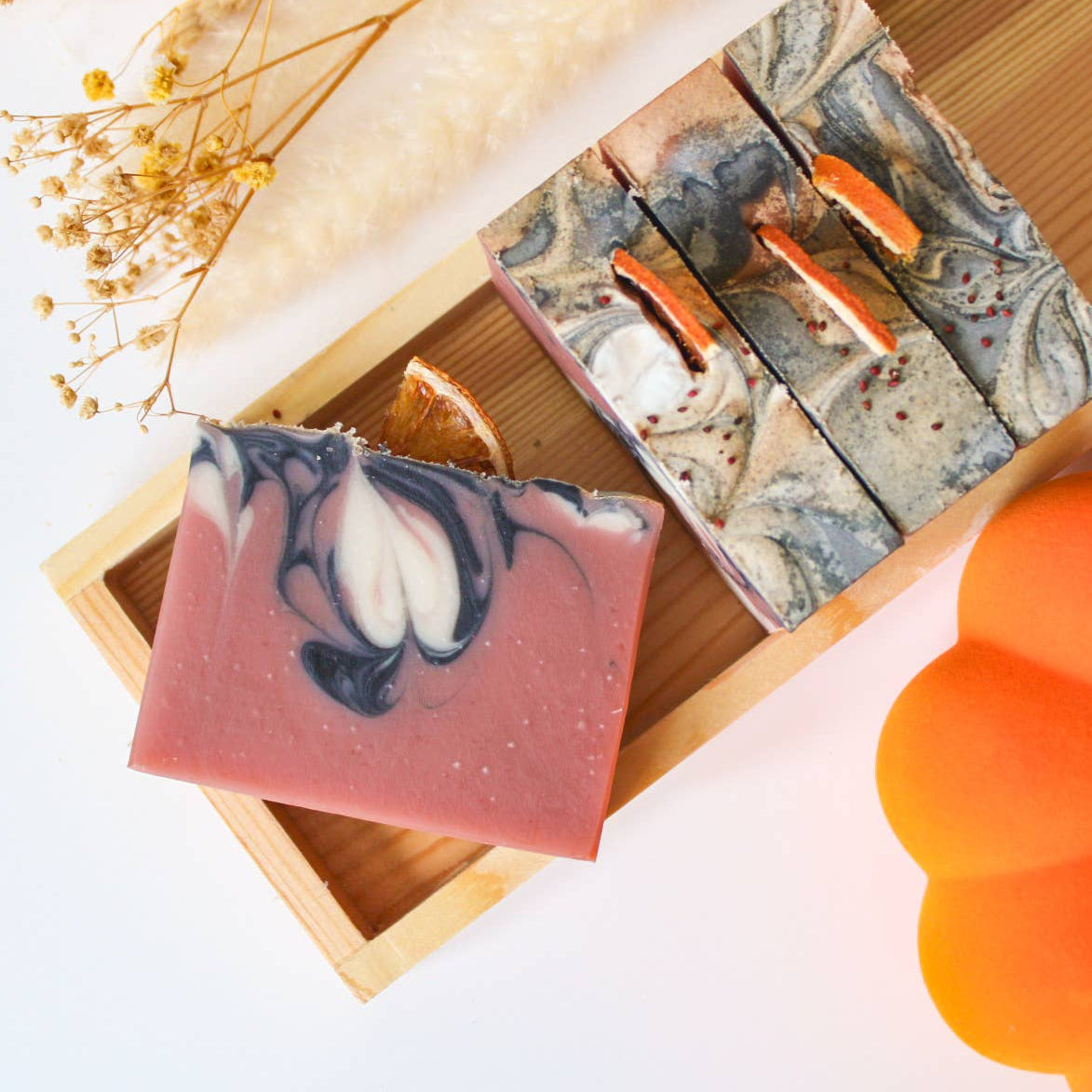 Three soap bars with natural designs on a wooden tray, surrounded by decorative elements including a pumpkin.