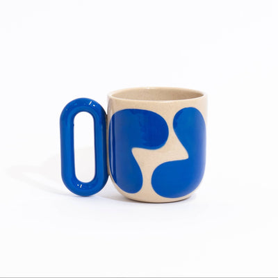 A ceramic coffee mug with a beige body, a bold blue abstract pattern, and a thick, matching blue handle, photographed on a seamless white background.