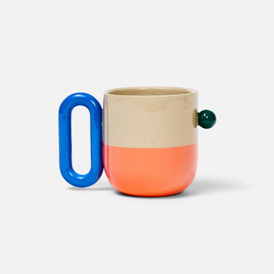 A ceramic mug with a split beige and coral body, a bright blue loop handle, and a small dark green spherical accent on the side, set against a plain white background.