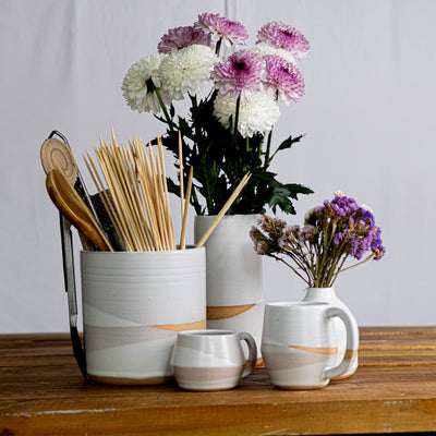 White ceramic containers with gold accents on a wooden surface, containing flowers and kitchen utensils.
