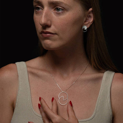 A woman with light brown hair wearing a silver circular pendant necklace and a matching silver stud earring, set against a solid black background.