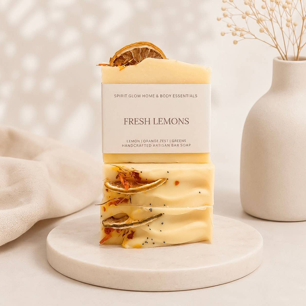 A bar of Fresh Lemons handcrafted artisan bar soap by Spirit Glow Home & Body Essentials, displayed on a white marble tray. The soap is pale yellow with dried orange slices and seeds, set against a warm, softly lit background with a beige cloth and a ceramic vase.