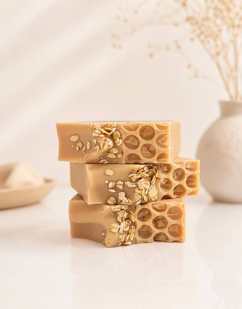 A bar of oatmeal and honey artisan soap displayed in two stacked pieces to show its honeycomb texture and oat topping, with a paper label on a warm, softly lit background.