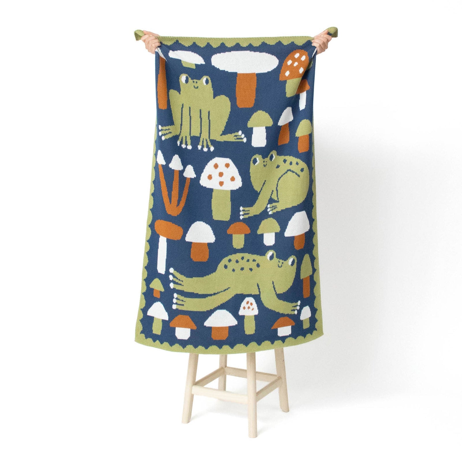 A rectangular woven blanket or throw, held up by two hands on a small wooden stand against a plain white background. The textile features a whimsical pattern of green frogs and orange and white mushrooms on a dark navy blue base, with a green scalloped border.