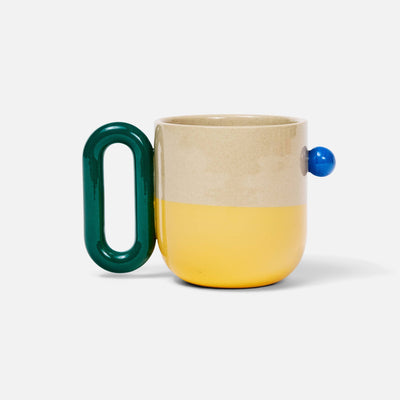 A ceramic mug with a split beige and yellow body, a dark green loop handle, and a small blue spherical accent on the side, set against a plain white background.
