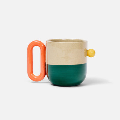 A ceramic mug with a split design, featuring a cream-colored upper half, a dark teal lower half, a bright coral handle, and a small yellow protruding accent on the side, set against a plain light gray background.