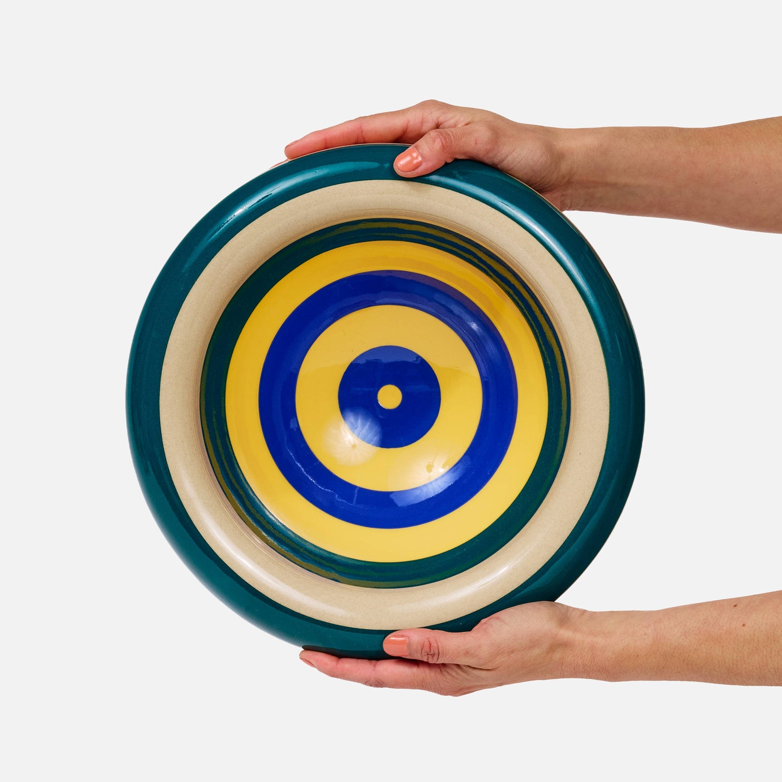 A person's hands holding a round, shallow dish with concentric target-style stripes in dark teal, cream, yellow, and blue against a plain white background.