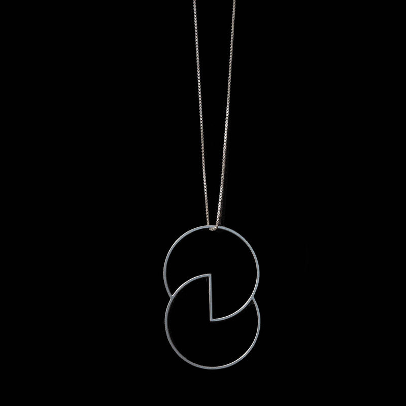 A close-up of a model wearing a delicate gold necklace with a large, open interlocking oval pendant. The model is wearing a light beige knit top against a solid black background.