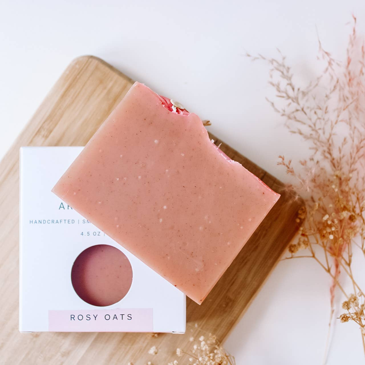 Pink soap bar on a wooden board with a label, surrounded by dried flowers on a light background