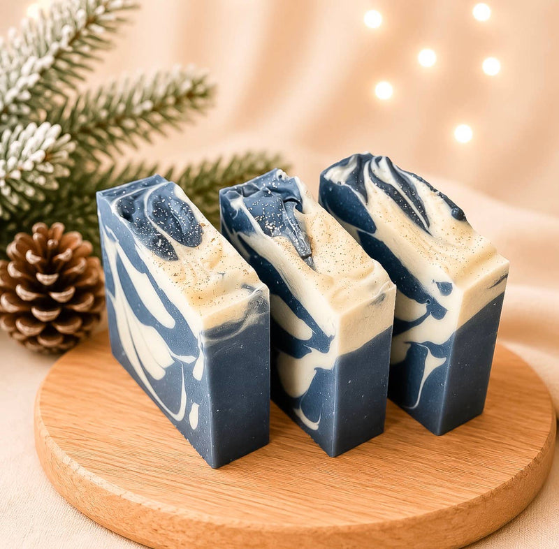 A handmade artisan bar soap labeled 'Nordic Lights' by Spirit Glow Home & Body Essentials, displayed on a round wooden board with a second soap piece showing its marbled blue and cream texture. The scene is decorated with a pine cone, evergreen branches, and warm bokeh string lights in the background.