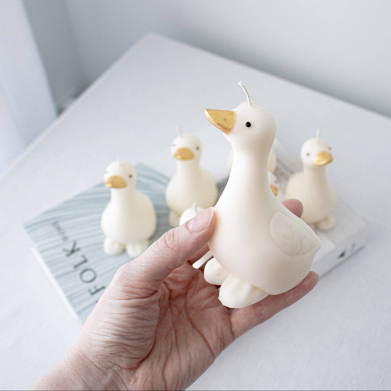 Six white goose shaped candles with gold beaks are arranged on top of a  book against a light gray background.