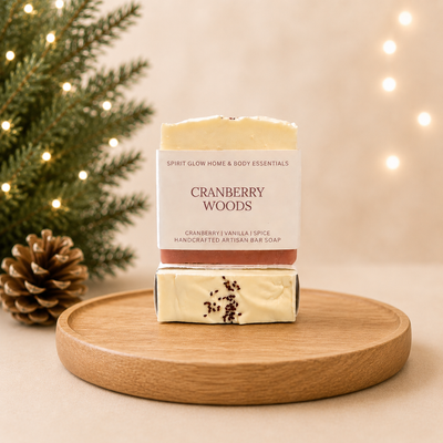 Two bars of Spirit Glow Handcrafted Artisan Bar Soap in the Cranberry Woods scent, stacked on a round wooden tray. The top bar is wrapped in a white paper sleeve with brown text, and the bottom bar is wrapped in a cream-colored paper sleeve decorated with dark cranberry sprigs.