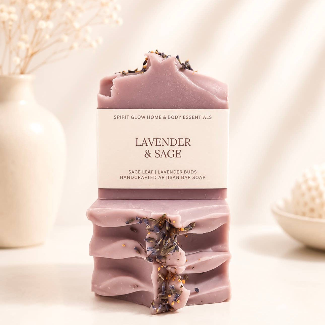 A handmade lavender and sage artisanal bar soap is displayed in a clean, styled setting with dried flowers in a white vase in the background. The soap is a dusty lavender-purple color with dried lavender buds embedded in the top and bottom, and features a white paper label with black text.