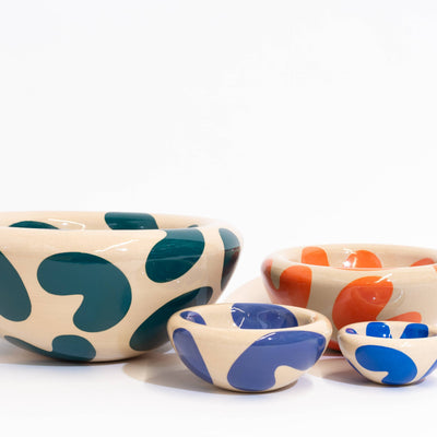 Four ceramic bowls in assorted sizes with abstract organic spot patterns in teal, orange, blue, and purple, arranged on a white studio background.