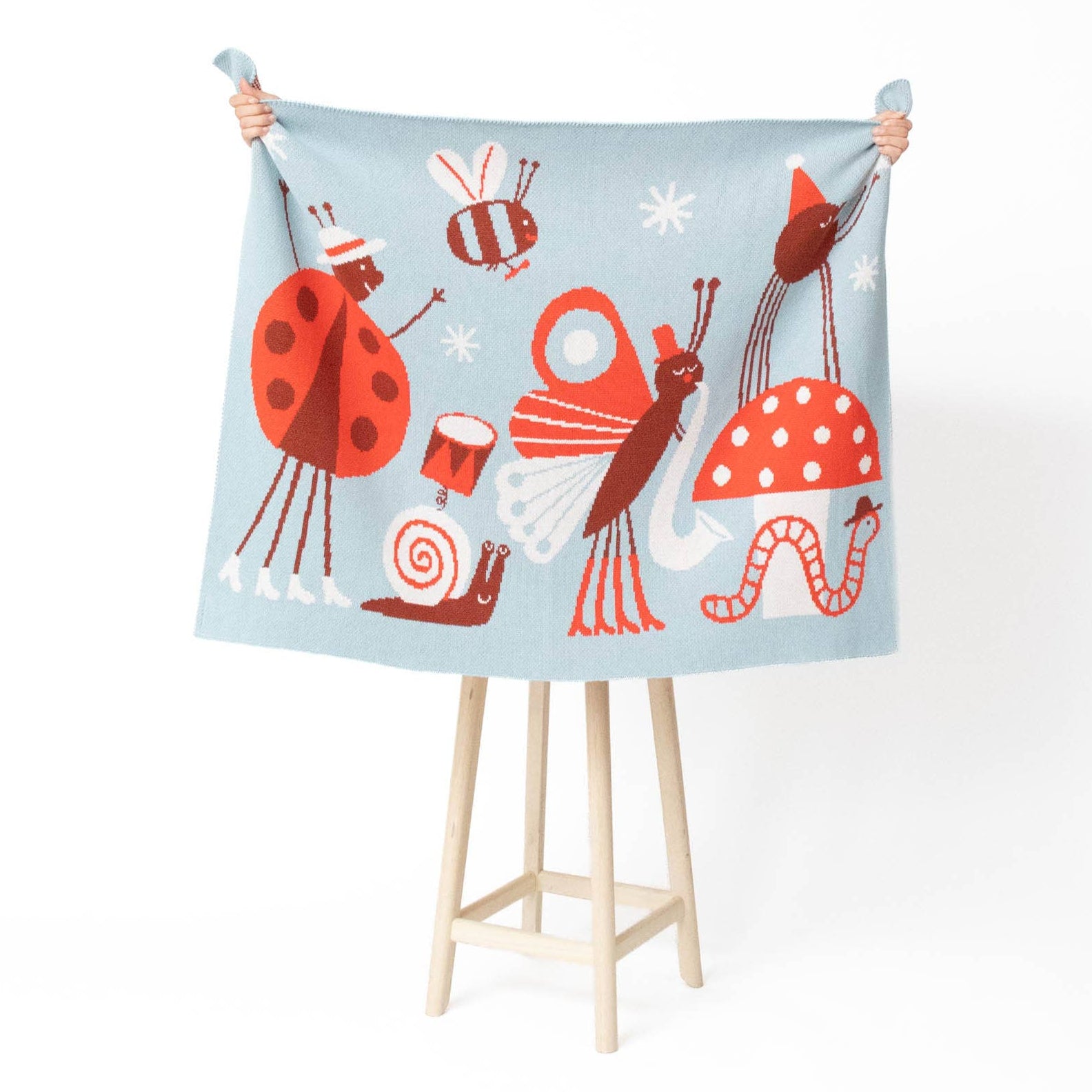 A light blue fabric blanket or throw featuring whimsical cartoon bugs and mushrooms, held up by a person's hands on a wooden stand against a white background.