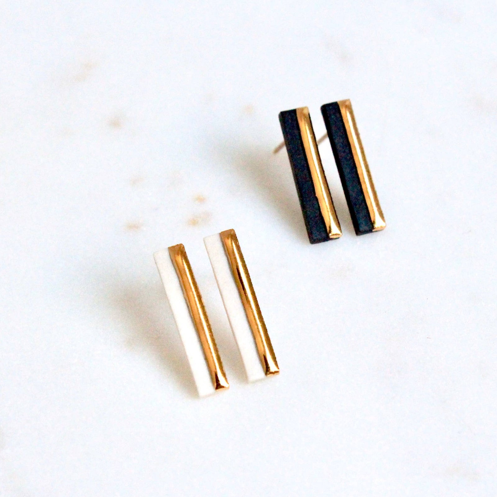 A pair of minimalist gold and  bar stud earrings resting on a light marble surface.