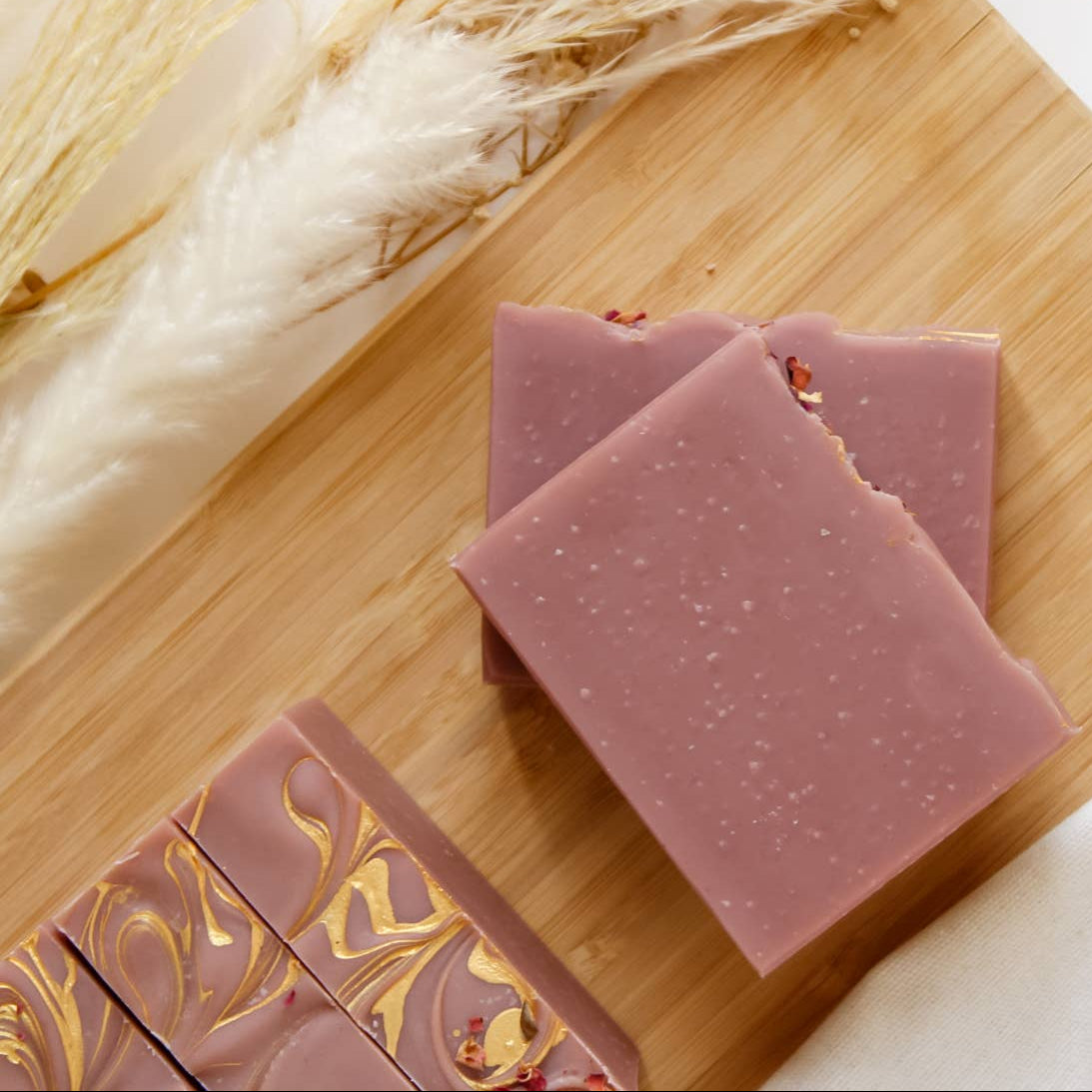 Soap bars with gold designs on a wooden board with dried plants and white fabric.