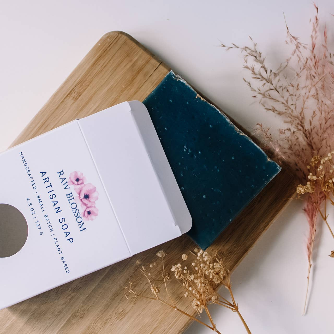 Artisan soap bar with packaging on a wooden board with dried flowers.