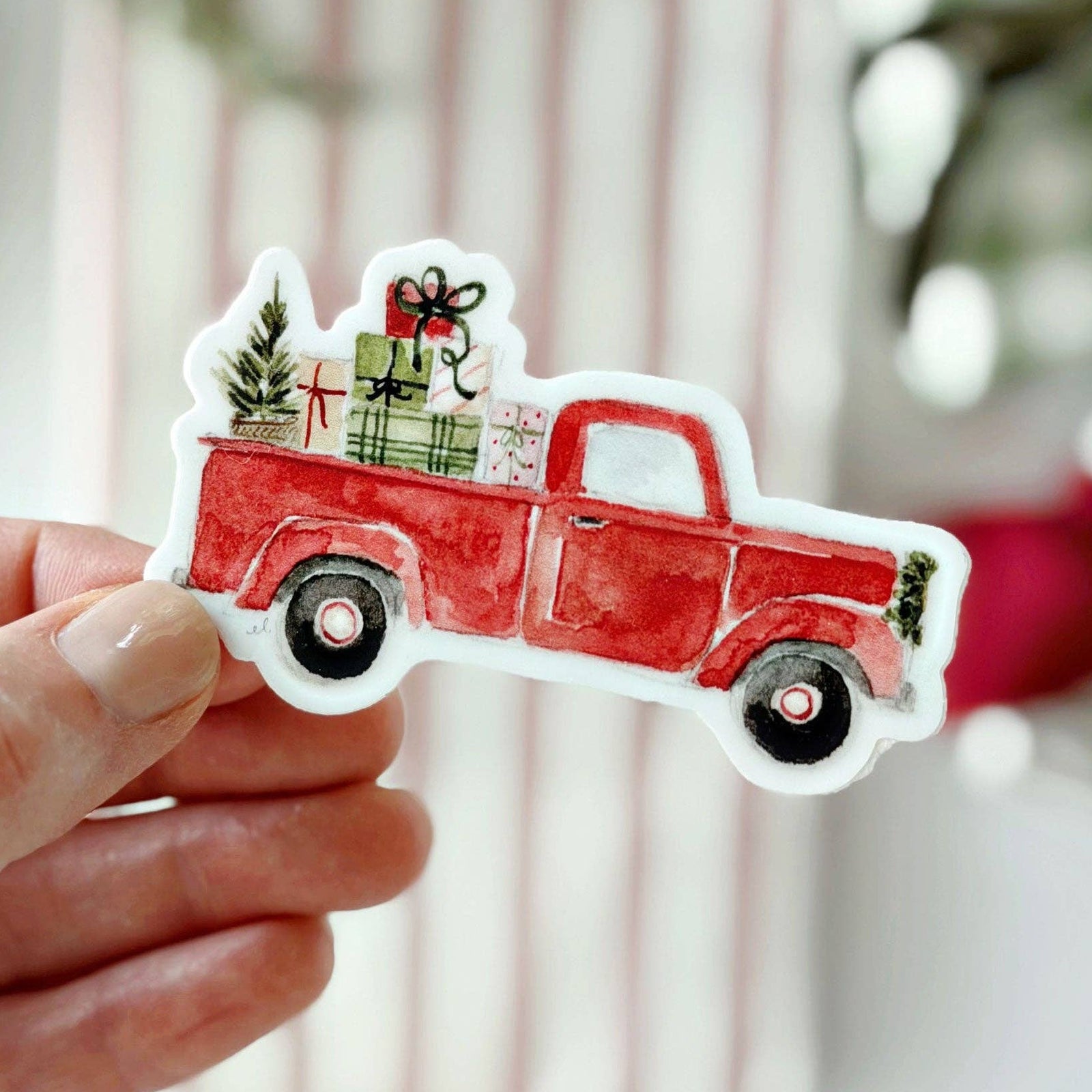 A hand holding a sticker of a vintage red Christmas truck on a light blurry background