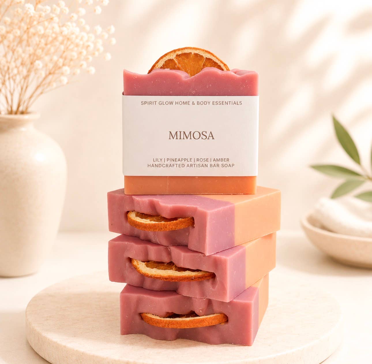 Four bars of Mimosa handcrafted artisan bar soap stacked on a round wooden tray. The soap bars are pink with a peach-colored side and a dried orange slice embedded in the top, set against a softly blurred background with a vase of dried flowers.