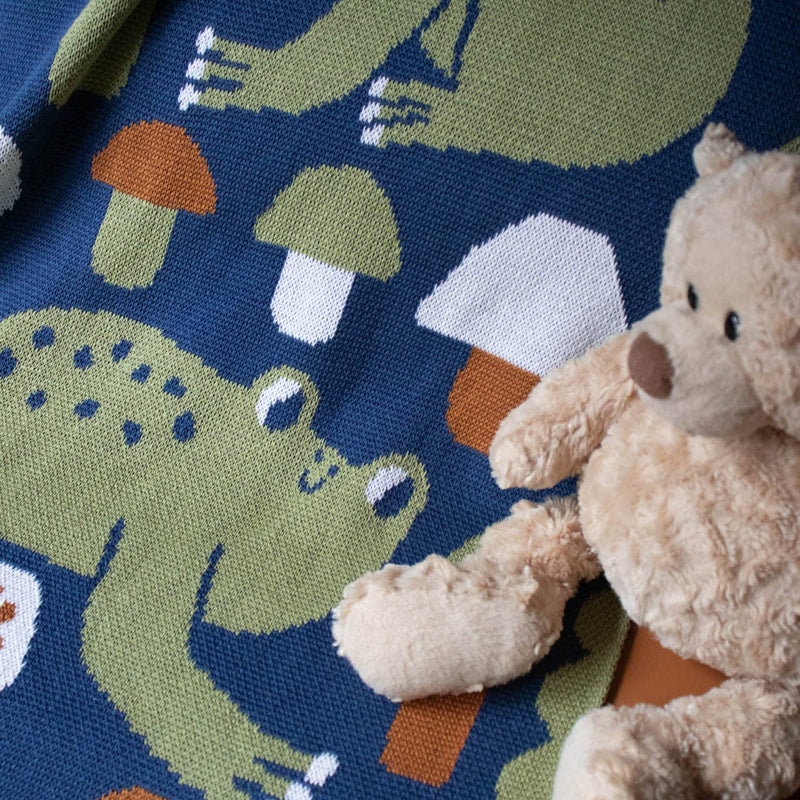 A rectangular woven blanket or throw, held up by two hands on a small wooden stand against a plain white background. The textile features a whimsical pattern of green frogs and orange and white mushrooms on a dark navy blue base, with a green scalloped border.
