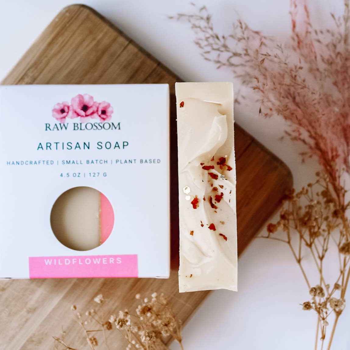 Raw Blossom Artisan Soap with packaging on a wooden board with dried flowers.